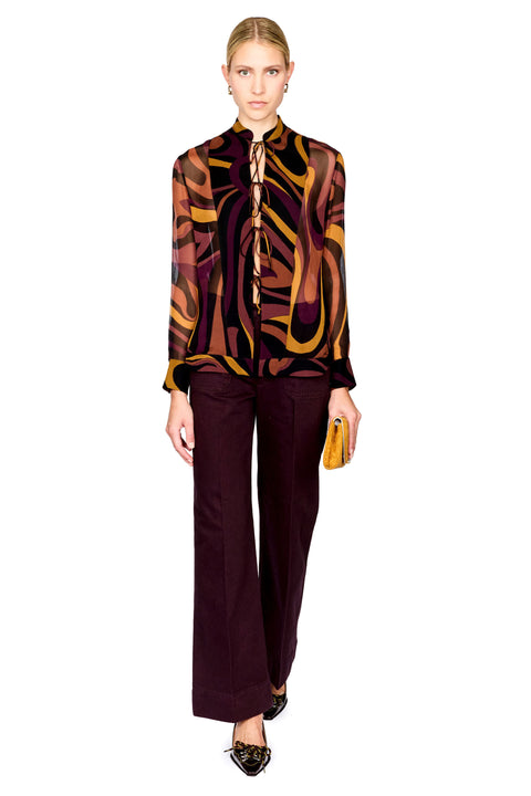 A full-length studio shot of a model wearing a long-sleeve silk shirt with an abstract wavy pattern in brown, gold, and black, paired with dark plum wide-leg trousers and holding a small yellow clutch.