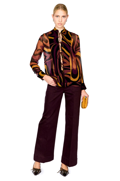 A full-length studio shot of a model wearing a long-sleeve silk shirt with an abstract wavy pattern in brown, gold, and black, paired with dark plum wide-leg trousers and holding a small yellow clutch.