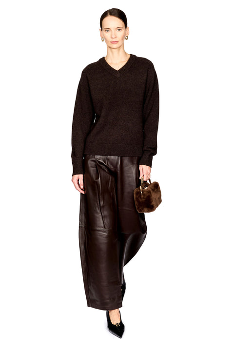 A model stands against a white studio background wearing a dark brown V-neck knit sweater, wide-leg dark brown faux-leather pants, black pointed shoes, and holding a small brown fur handbag.