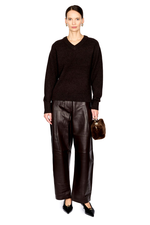 A model stands against a white studio background wearing a dark brown knit V-neck sweater, dark brown wide-leg leather pants, black pointed flats, and holding a small dark brown fur-like handbag.