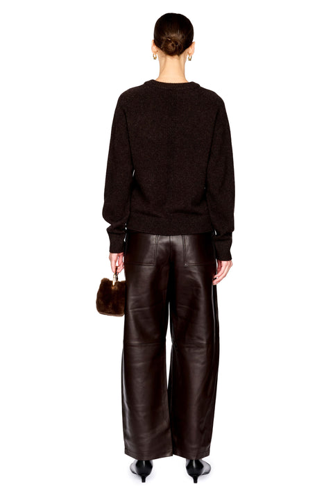 A model shown from the back wearing a dark brown knit sweater and wide-leg dark brown leather pants, holding a small brown fur accessory against a plain white background.