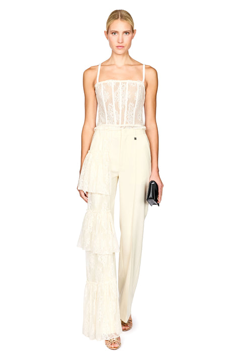 A model wearing a cream-colored two-piece outfit consisting of a lace camisole top and matching high-waisted wide-leg trousers with a ruffled tulle overlay, holding a small black clutch against a plain white background.