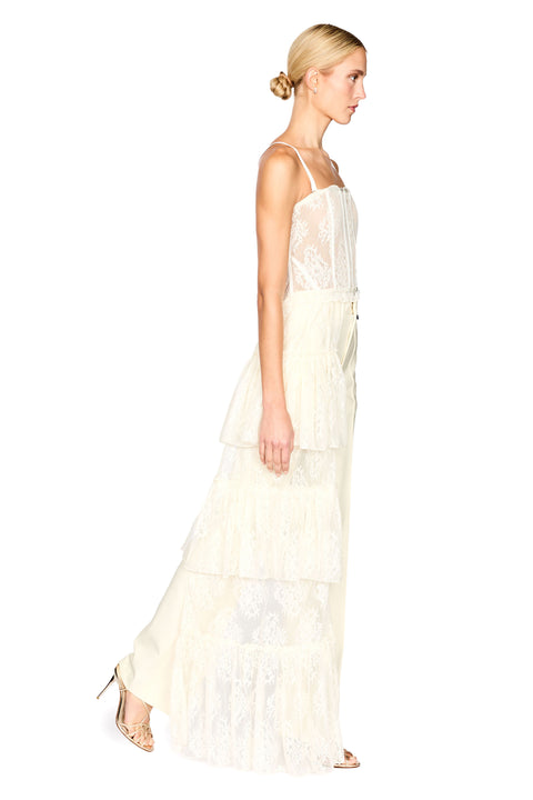 A woman modeling a long cream lace top with thin straps and a tiered skirt, shown in profile against a plain white studio background.