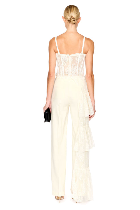 A woman shown from the back wearing a cream-colored lace and mesh top with thin straps and matching pants, a structured lace bodice, and layered ruffle details on the pants. She is holding a small black clutch against a solid white background.