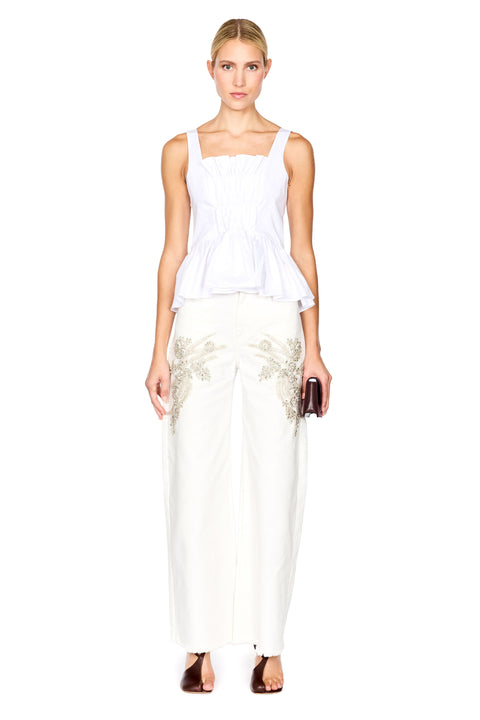A woman modeling a white peplum top and white wide-leg trousers with gold floral embroidery, holding a small dark clutch against a plain white background.