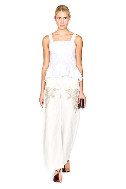 A woman modeling a white peplum top and white wide-leg trousers with gold floral embroidery, holding a small dark clutch against a plain white background.
