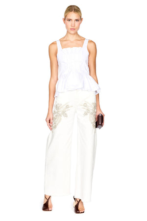 A woman modeling a white peplum top and white wide-leg trousers with gold floral embroidery, holding a small dark clutch against a plain white background.