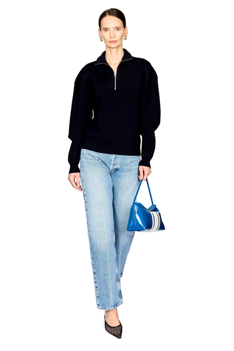 A woman modeling a navy long-sleeve quarter-zip sweater, light-wash blue jeans, and black pointed shoes, while holding a shiny blue handbag with white stripes against a plain white background.
