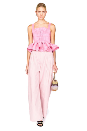 A model wearing a pink sleeveless top with a gathered peplum hem and matching wide-leg trousers, holding a small striped handbag against a plain white background.