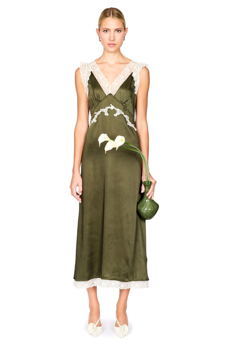 A woman models a sleeveless olive green satin maxi dress with white lace trim at the V-neckline, waist, and hem. She is holding a small green handbag and wearing white pointed-toe flats, set against a plain white studio background.