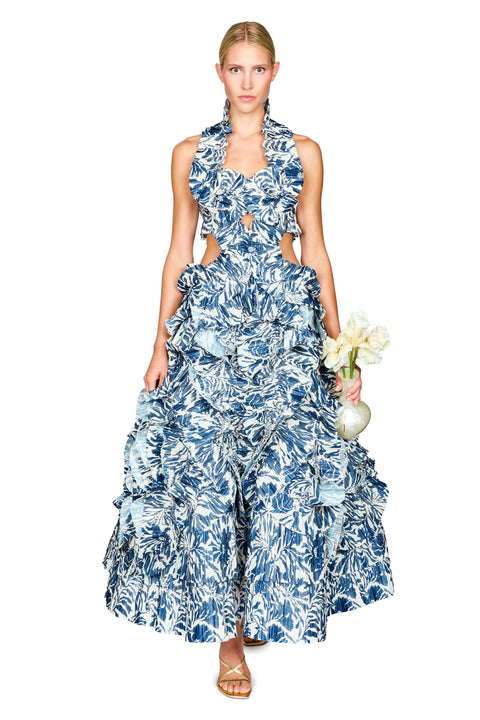 A model wearing a sleeveless, tiered maxi dress with a blue and white tropical floral print, holding a small white vase clutch with flowers against a solid white background.