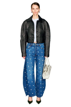 A model stands against a white background wearing a black leather jacket, blue wide-leg jeans with white polka dots, a white top, and carrying a white handbag.