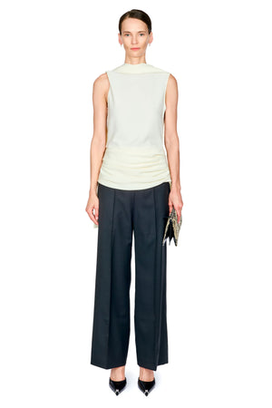 A model stands against a plain white background wearing a sleeveless white mock-neck top, dark black wide-leg trousers with white waist ruching, and black pointed heels, while holding a small patterned clutch.