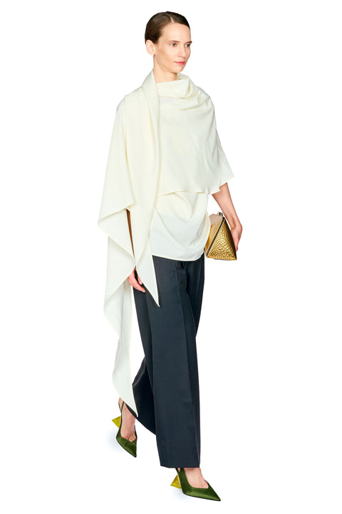 A model is shown from the side wearing a cream draped wrap top, dark black wide-leg trousers, olive green pointed-toe heels, and holding a small gold woven clutch against a solid white background.