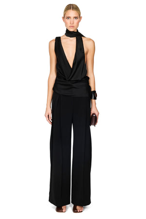 A model wearing a black satin halter-neck top with a draped neckline and matching wide-leg trousers, styled with a black choker, black cuff bracelet, and holding a small dark clutch against a plain white background.