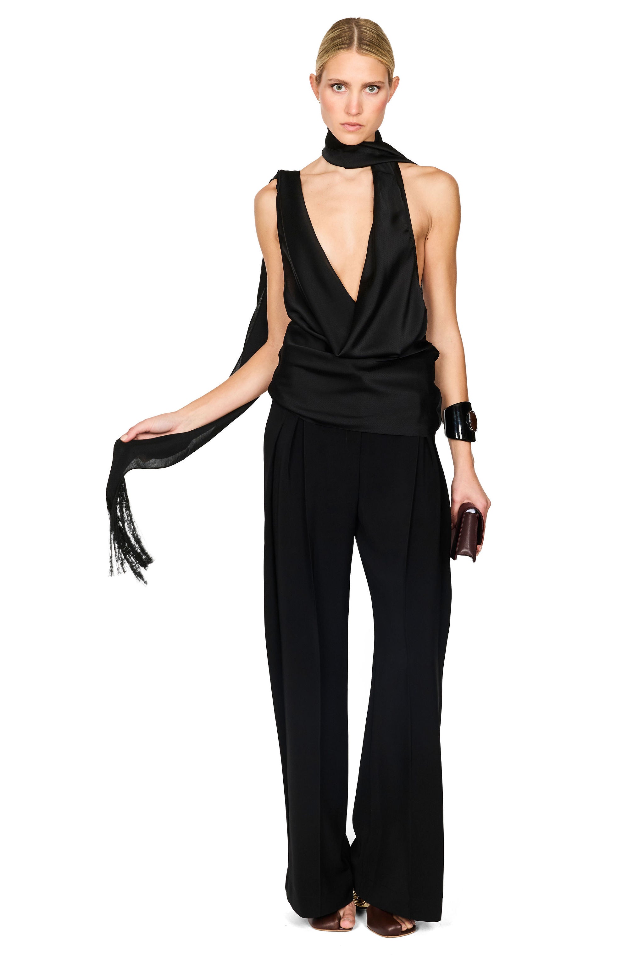 A model wearing a black sleeveless wrap-style top with a deep V-neck, wide-leg pants, and a matching draped scarf, standing against a plain white background.