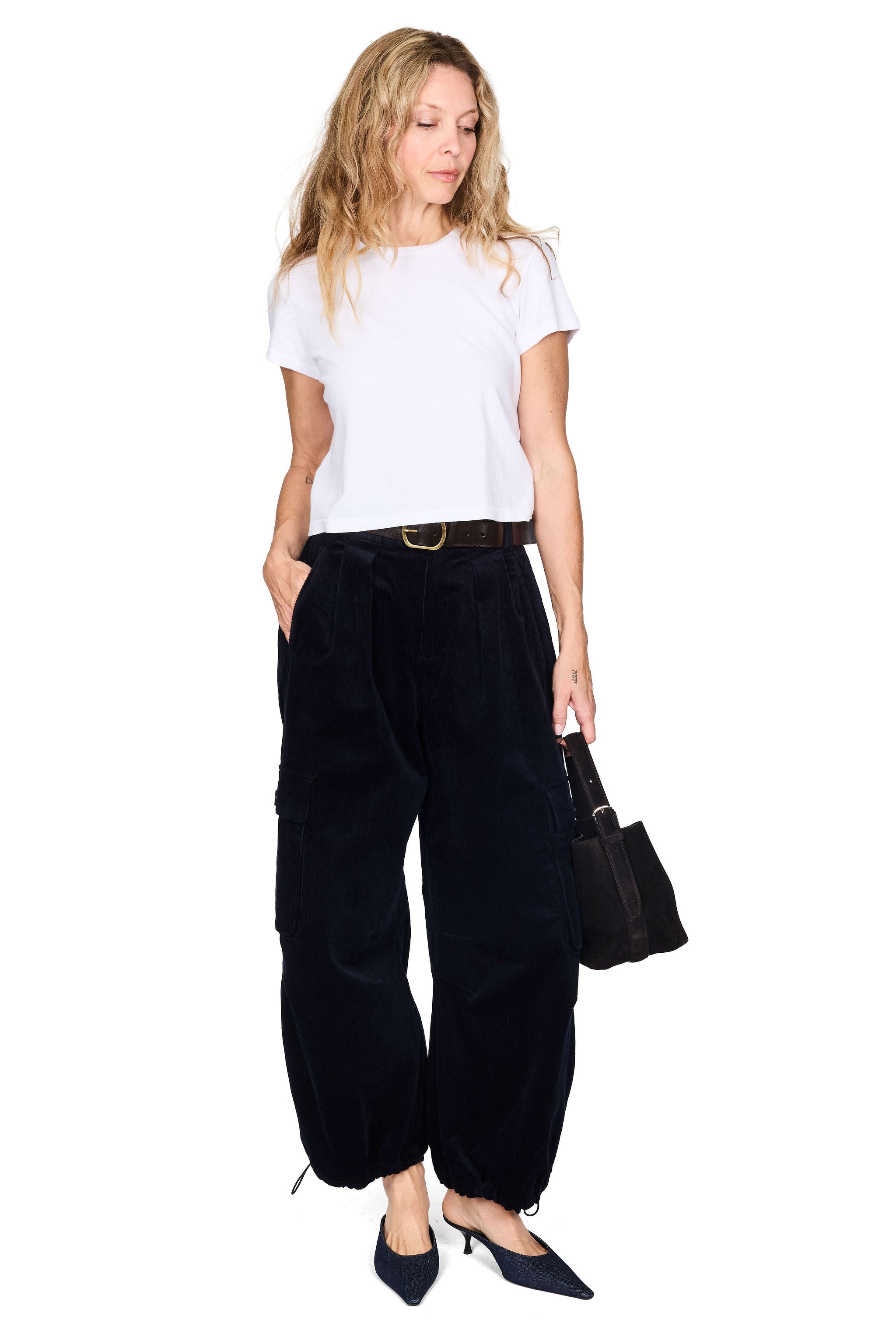 A woman modeling black wide-leg velvet cargo pants with a dark brown belt, paired with a white t-shirt, black pointed-toe heels, and holding a black handbag against a plain white background.
