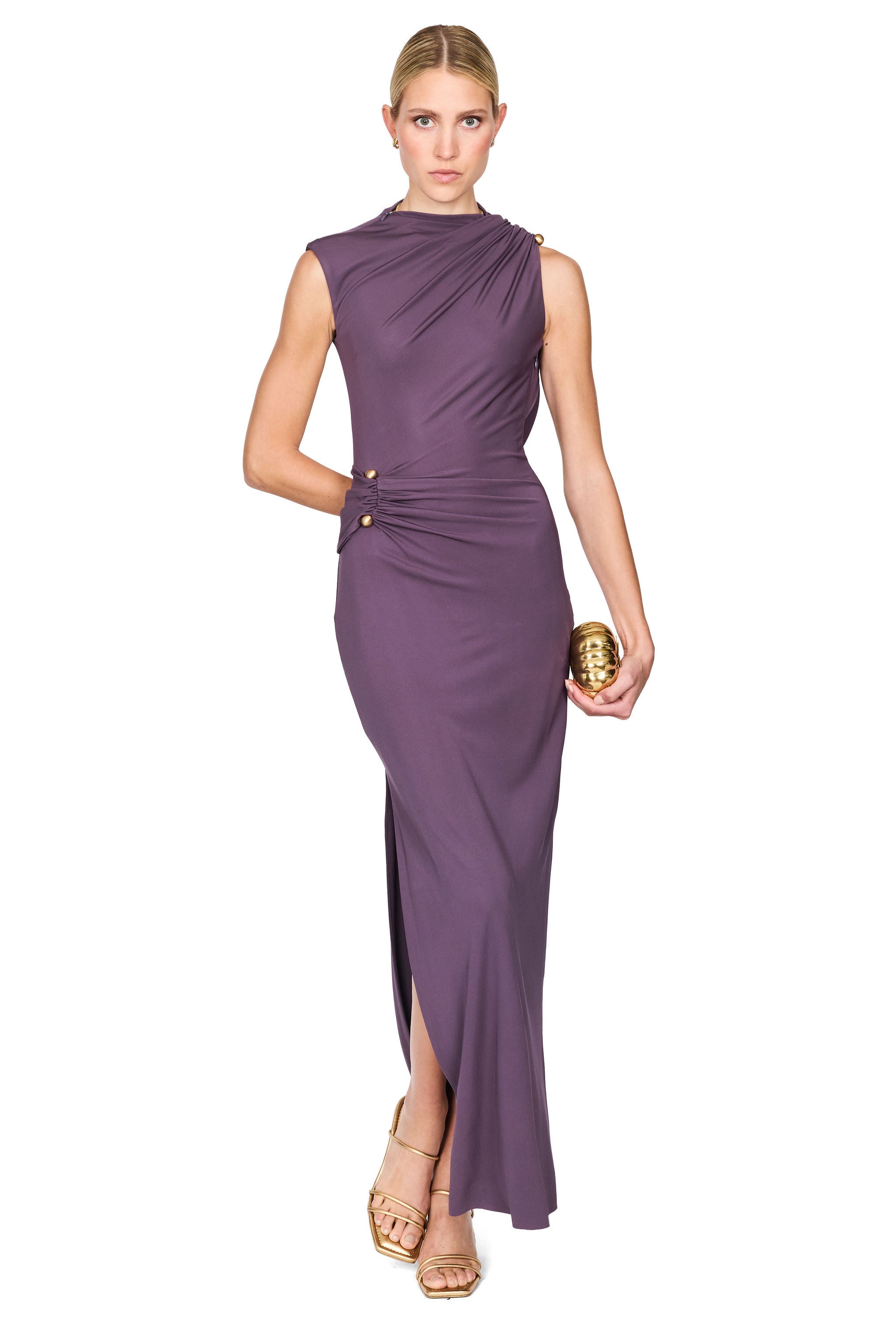 A woman modeling a sleeveless, draped floor-length gown in a muted purple/plum color, paired with metallic gold sandals and holding a small gold clutch, set against a plain white background.