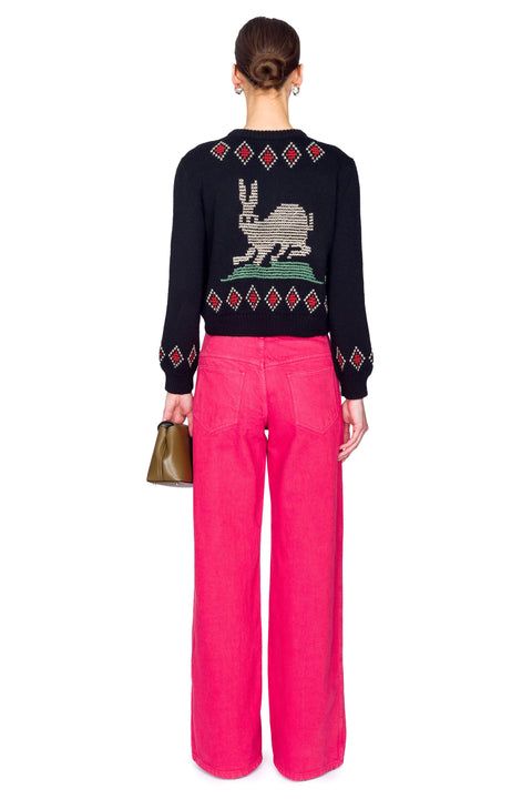 Woman wearing a black embroidered cardigan and pink pants on a white background from the back.