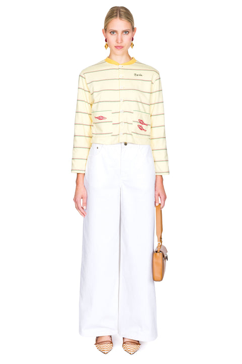 Woman wearing a yellow striped sweater with red fish on the pockets and white pants on a white background with one hand clutching bag.