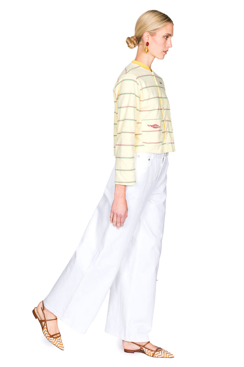 Woman wearing a yellow striped sweater with red fish on the pockets and white pants on a white background from a side view.