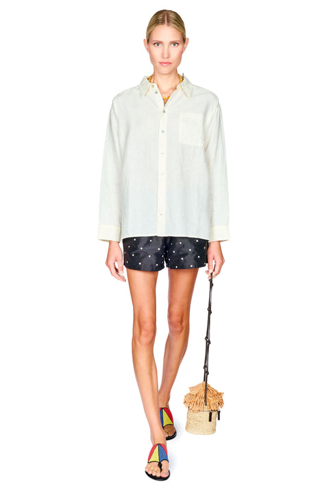 A model stands against a white background wearing a light cream long-sleeve button-up shirt, black shorts with white dot patterns, and colorful sandals, while holding a small woven straw bag with a dark strap.