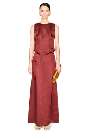 A model stands against a white background wearing a sleeveless, floor-length rust-red set. She is holding a small gold clutch and wearing light-colored sandals.