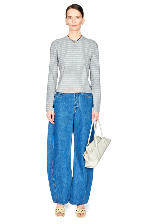 A model stands against a white background wearing a gray and white striped long-sleeve top, wide-leg blue jeans, beige sandals, and holding a light beige handbag.