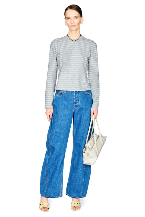 A model stands against a white studio background wearing a gray and white horizontally striped long-sleeve knit top, blue wide-leg denim jeans, and holding a light beige tote bag.