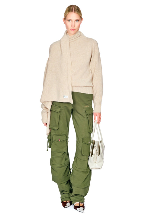 A model stands against a white background wearing a beige mock-neck knit top, olive green cargo pants, and holding a white tote bag.