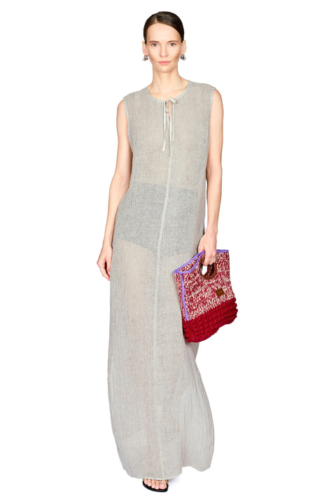 A model wearing a sleeveless, light gray linen maxi dress with a tie-neck detail, holding a red and purple patterned woven clutch bag against a plain white background.