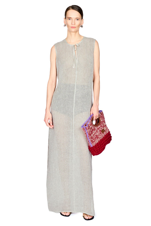 A model wearing a sleeveless, light gray linen maxi dress with a tie-neck detail, holding a red and purple patterned woven clutch bag against a plain white background.