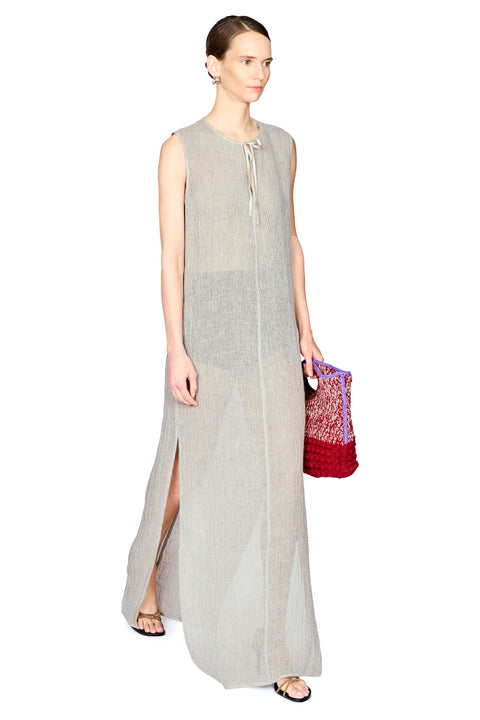 A woman modeling a sleeveless, floor-length light gray linen dress with a tie-neck detail and a side slit, holding a red and purple patterned woven clutch bag against a plain white background.