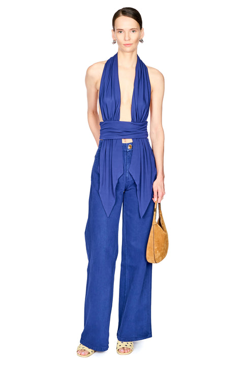 A model wearing a royal blue halter-neck top with wide-leg pants, holding a tan leather handbag against a plain white background.