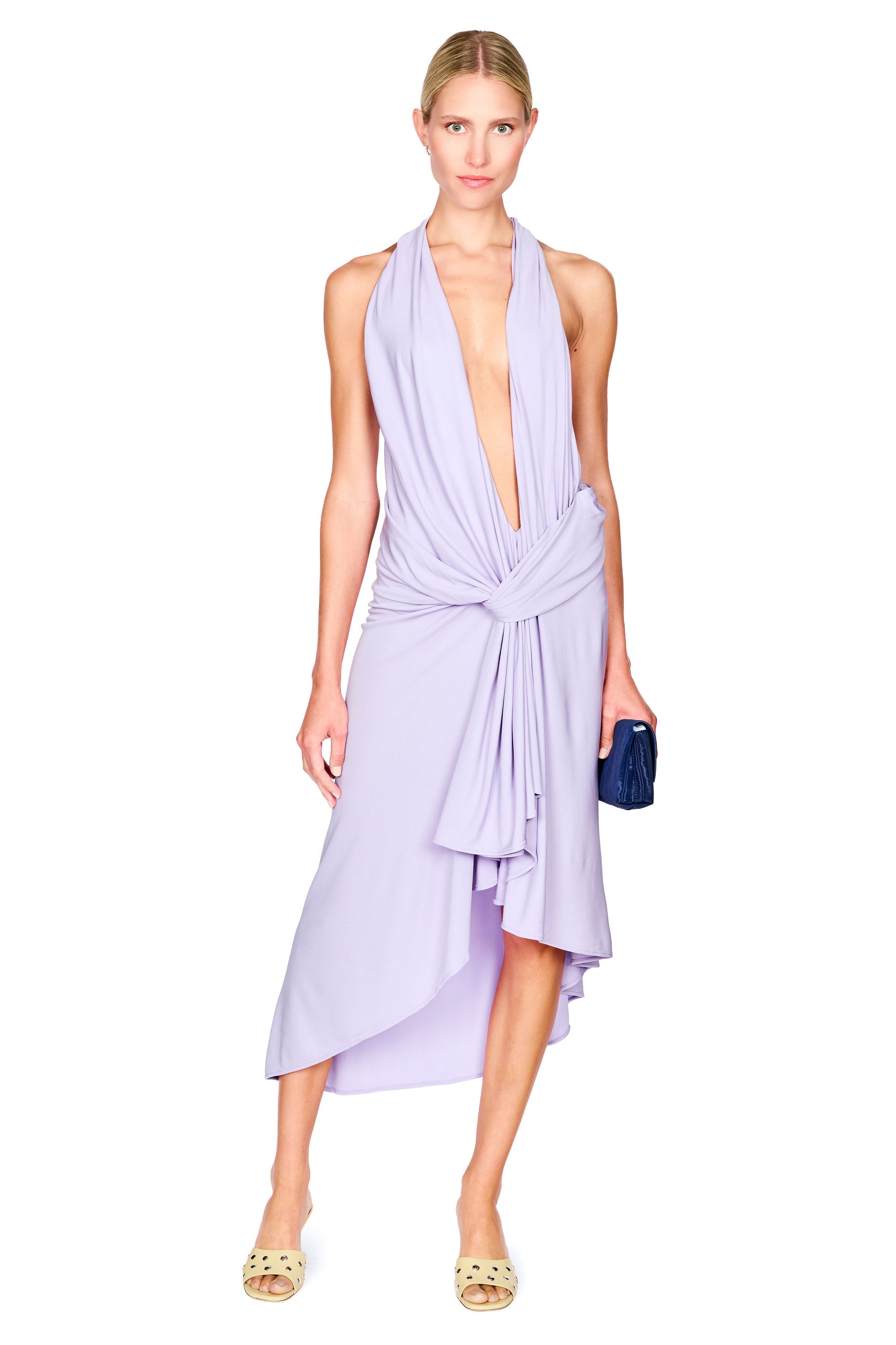 A model wearing a lavender halter wrap midi dress with a deep plunging V-neckline and a knotted waist, paired with gold sandals and holding a small navy blue clutch, set against a plain white background.