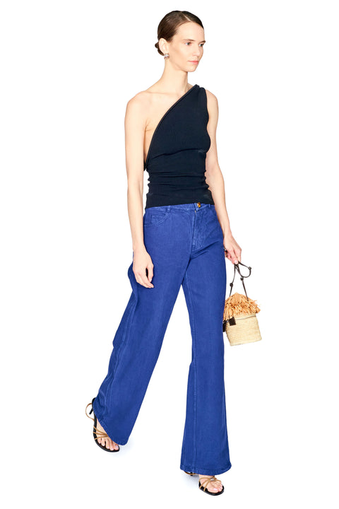 A model stands against a white background wearing a black one-shoulder top, blue wide-leg jeans, and holding a small beige woven handbag.