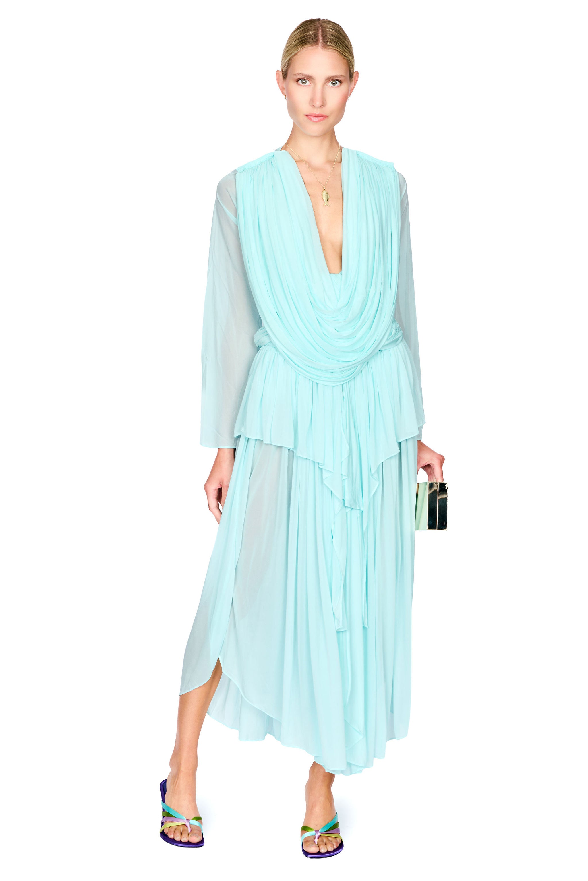 A model wearing a light aqua blue pleated long-sleeve top and maxi skirt set with a draped V-neckline, paired with colorful flat sandals and holding a black clutch, set against a plain white background.