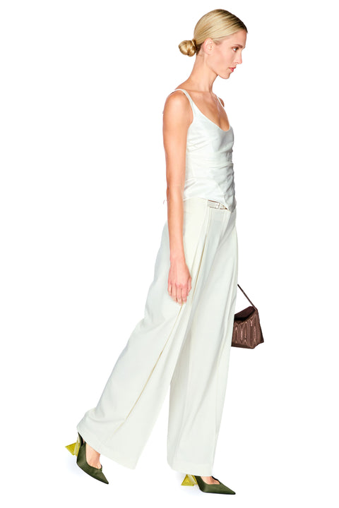 A woman in profile wearing a light cream satin camisole and matching wide-leg trousers, styled with olive green pointed shoes and a small brown handbag against a plain white background.