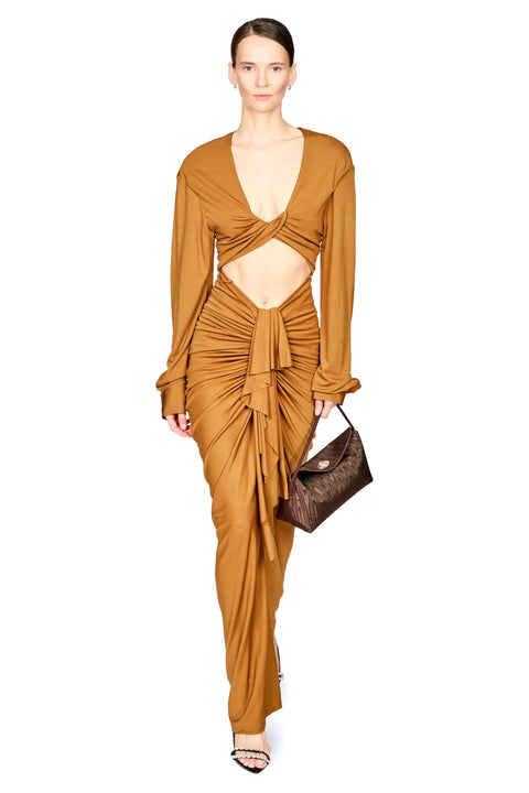 A woman on a white background wearing a cognac brown long dress with long sleeves, drapery from the waist down, and a cutout under the chest