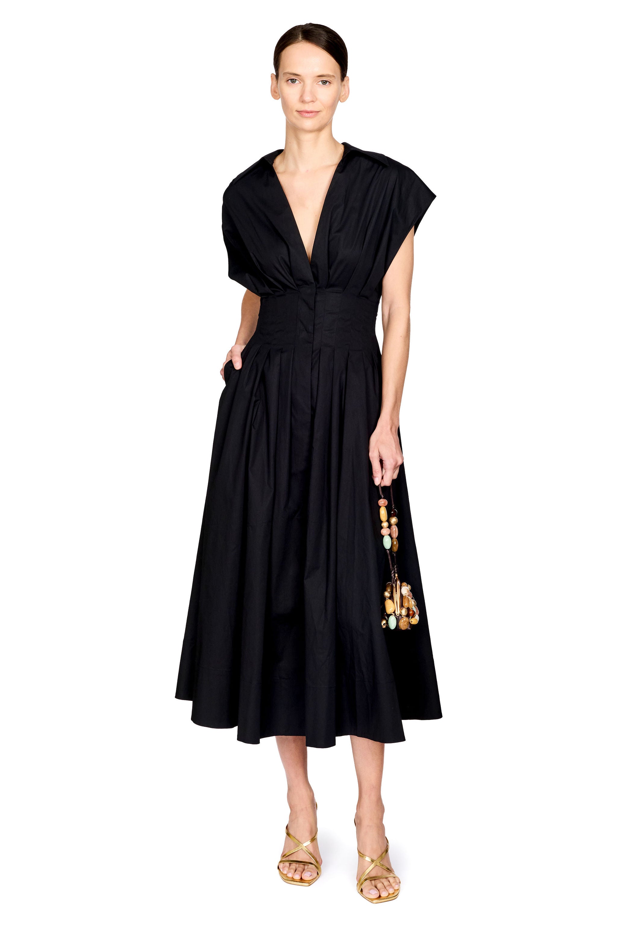 A model wearing a black midi dress with a deep V-neckline, cap sleeves, and a gathered waist, paired with gold sandals and a beaded bag, set against a plain white background.