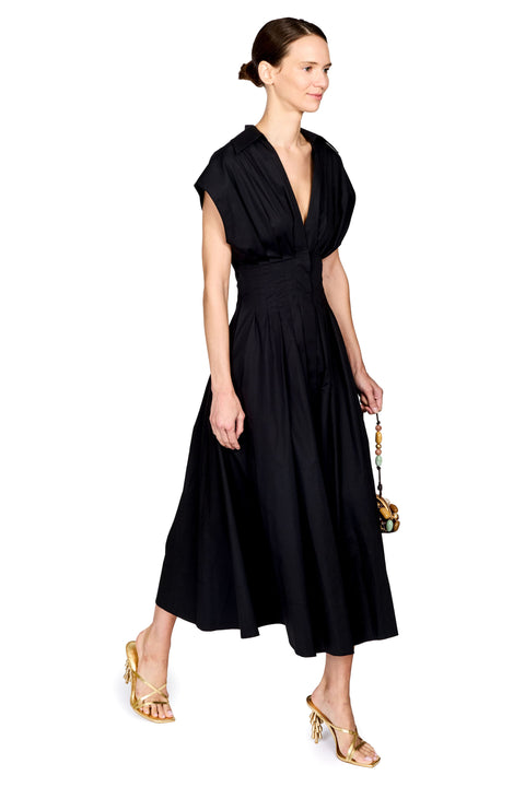 A woman modeling a black midi dress with a deep V-neckline, cap sleeves, and a gathered waist, paired with gold heeled sandals and a small gold handbag, set against a plain white background.