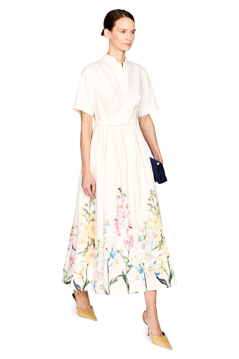 A woman modeling a cream-colored midi dress with a high collar, short sleeves, and a colorful floral print along the hem, paired with yellow pointed-toe heels and holding a small dark blue clutch, set against a plain white background.