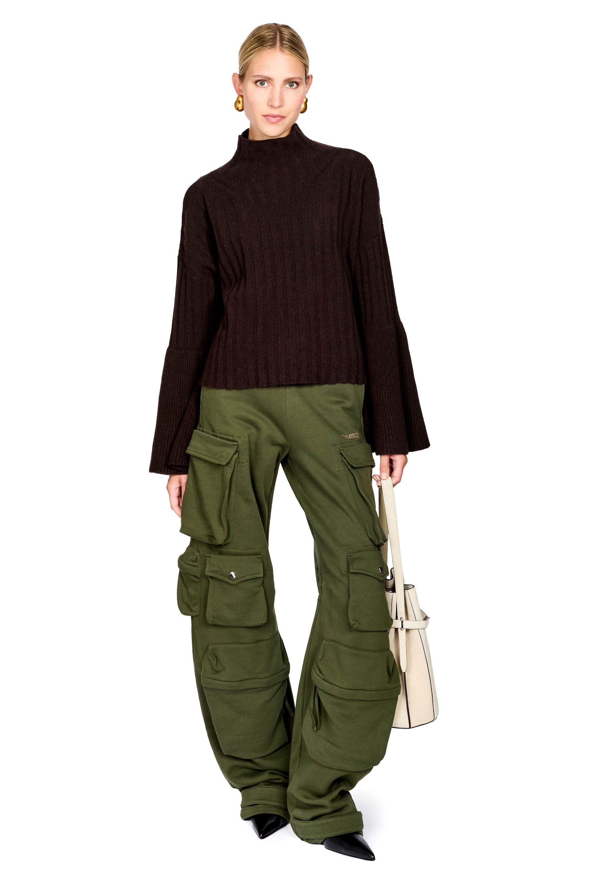 A model stands against a white background wearing a dark brown ribbed mock-neck sweater, olive green cargo pants, and holding a light beige shoulder bag.
