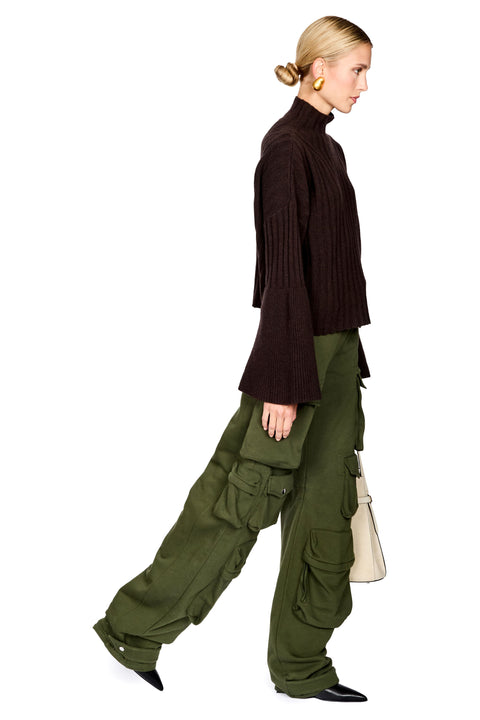 A side-profile studio shot of a model wearing a dark brown ribbed turtleneck sweater, olive green cargo pants, and black pointed shoes, holding a light beige handbag against a plain white background.