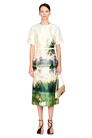 A model stands against a white background wearing a short-sleeve midi dress with a cream-colored textured top and a green and blue landscape print featuring a swan. She is holding a beige handbag and wearing brown strappy heels.