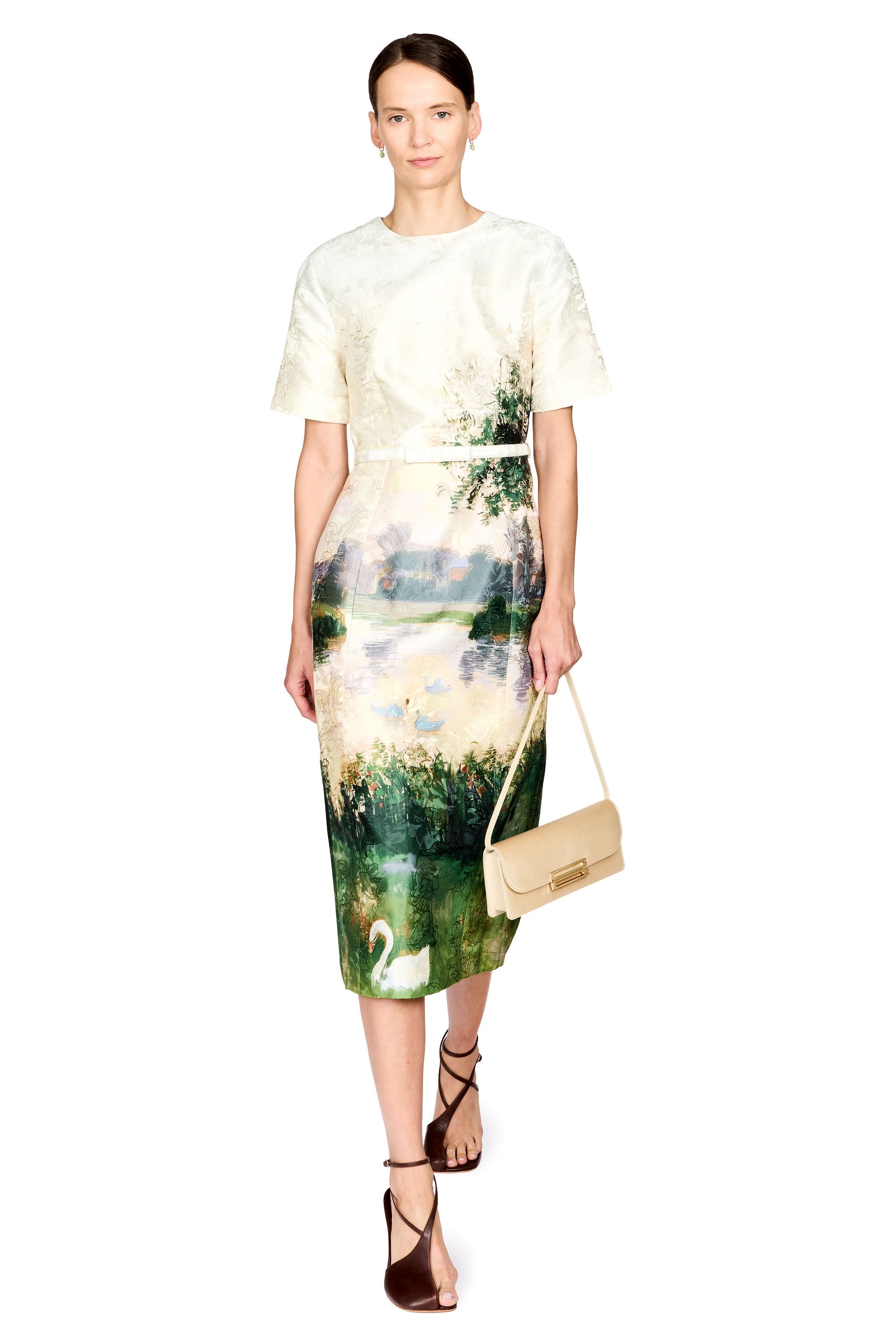 A model wearing a short-sleeve midi dress with a cream lace bodice and a watercolor landscape skirt featuring green trees, a lake, and a white swan. She is holding a beige shoulder bag and wearing brown strappy heels against a white background.