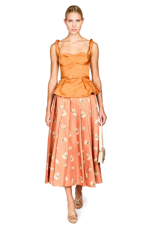 A model wearing a two-piece outfit consisting of a solid orange peplum top and a matching orange midi skirt with a white floral print, paired with beige high heels and holding a small beige handbag against a plain white background.