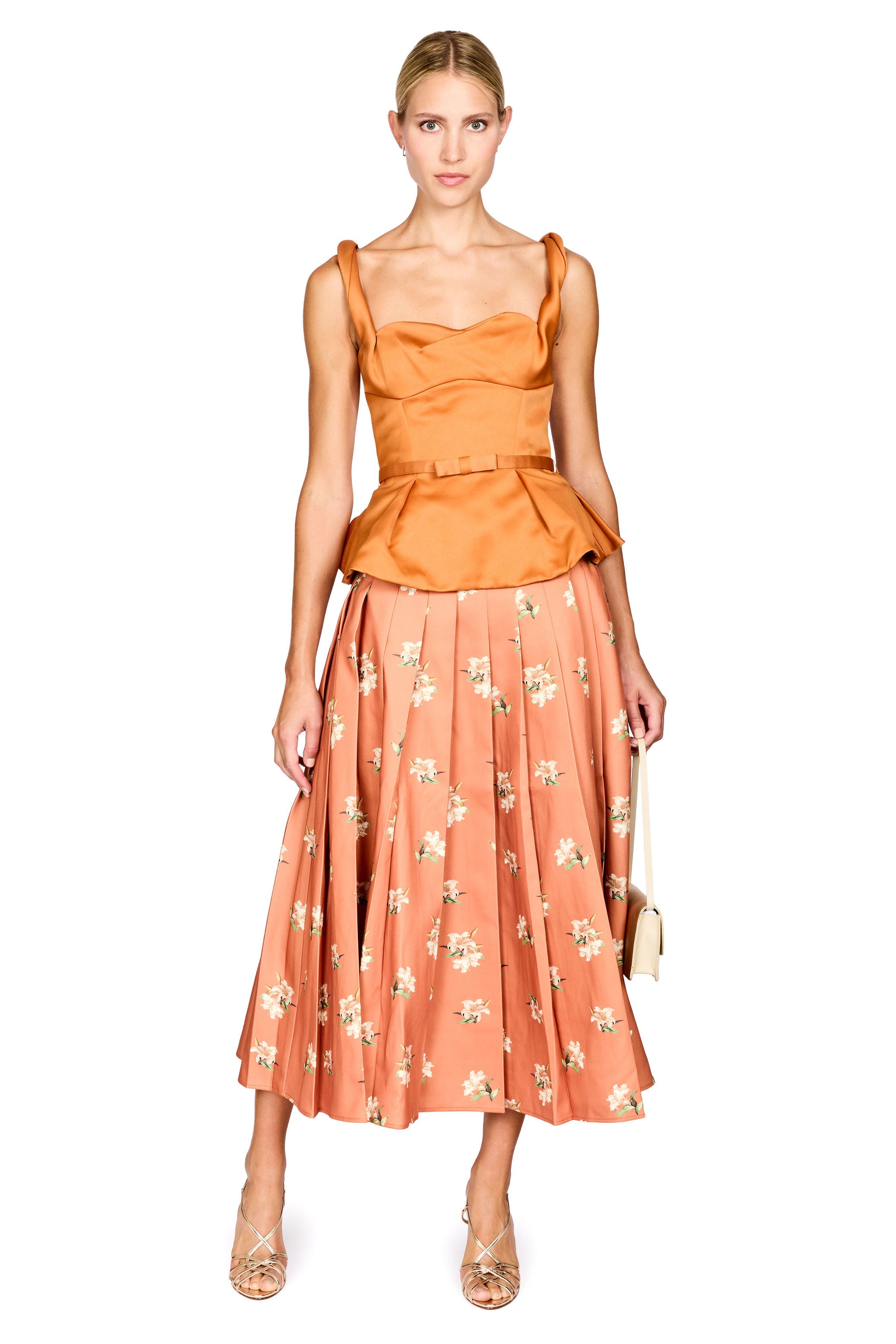 A model wearing a two-piece outfit consisting of a cropped, satin-like orange-brown top with shoulder straps and a peplum waist, paired with a pleated midi skirt in a peachy-orange color with a small white floral pattern. She is holding a small beige handbag and wearing metallic gold strappy heels against a plain white background.