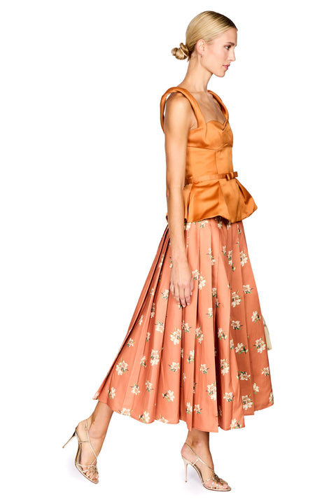 A woman modeling a sleeveless peach-orange top with a peplum waist and a matching floral midi skirt, paired with clear high-heeled sandals, against a plain white background.