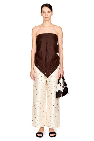 A model stands against a white studio background wearing a strapless dark brown top with a draped, asymmetrical hem and matching dark polka-dot wide-leg pants. She is holding a black and white patterned handbag and wearing black slide sandals.